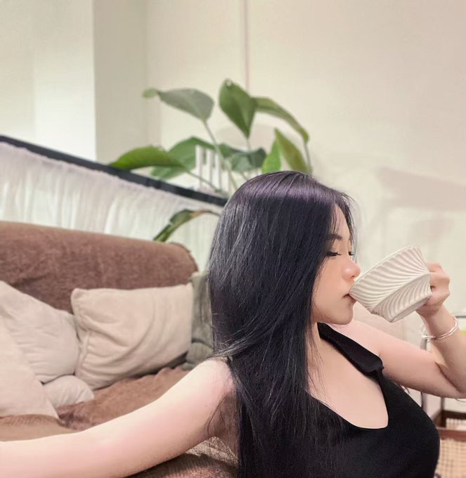 A young woman with long black hair sips from a patterned white mug while relaxing in a cozy living room with a brown sofa and green plant. The image pairs with a Mid-Autumn Festival promotional post offering an exclusive red envelope code "GATE-r2LFHe" with Gate, though no text or branding is visible in the photo itself.
