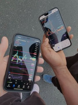 Two people holding smartphones displaying stock trading charts with red and green candlesticks on a paved outdoor surface, comparing their trading apps side by side.