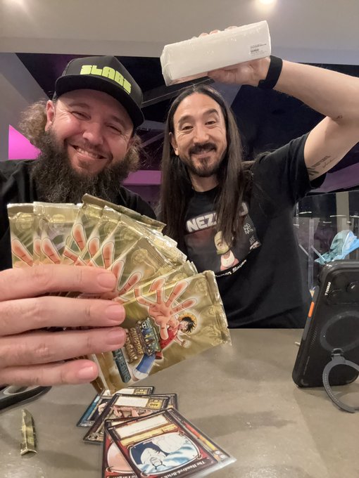 Two smiling men at what appears to be a trading card event, one holding a fan of gold 'Topps' cards and another holding up a silver graded card slab.