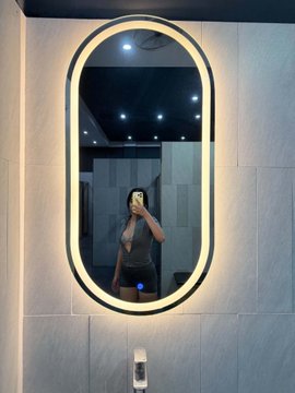 A person takes a selfie in an oval-shaped, illuminated LED bathroom mirror mounted on a tiled wall, with a soap dispenser visible below.