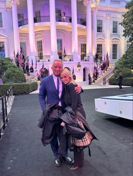 A man and woman pose together outside the White House, which is lit up with pink and purple lighting at night.