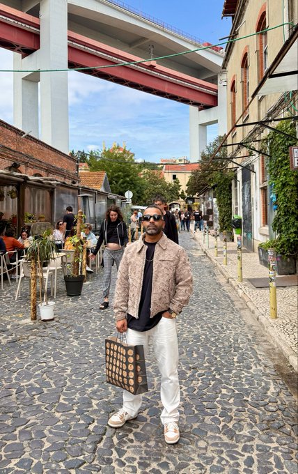 A man wearing a beige patterned jacket, white pants, and sunglasses stands on a cobblestone street holding a black and white polka-dot shopping bag.
