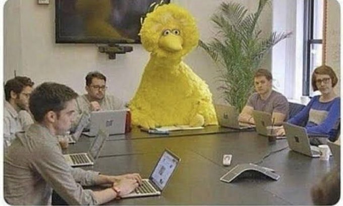 Big Bird from Sesame Street is seated at the head of a corporate conference table surrounded by professionals working on laptops during a business meeting.