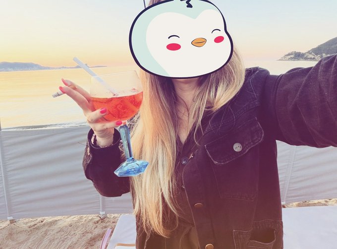 A person with long blonde hair holding a pink cocktail and a cigarette at sunset by the sea, with their face obscured by a cute cartoon sticker.