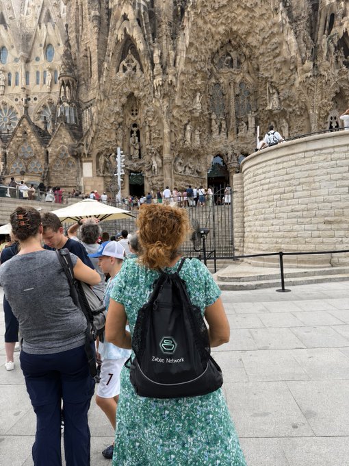 A woman with curly hair stands with her back to the camera in front of the intricate stone façade of Barcelona's Sagrada Família, wearing a backpack featuring the "Zebec Network" logo, connecting the travel moment to the $ZBCN cryptocurrency.