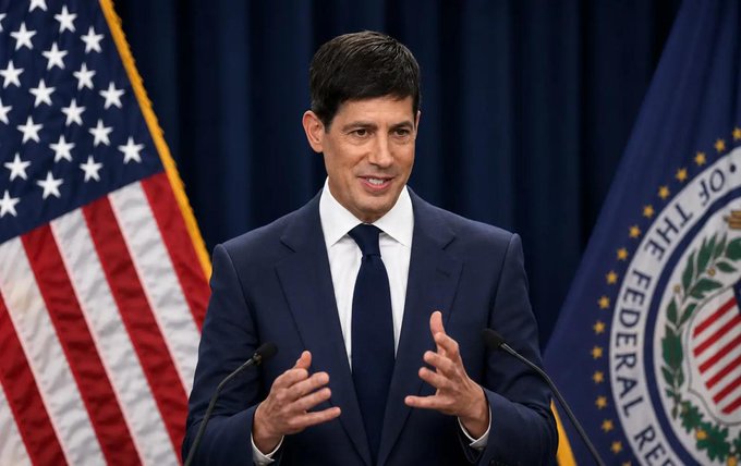 The image shows a man standing at a podium with his hands clasped behind his back, suggesting he may be giving a speech or presentation. The background features two flags, one representing the United States and the other the Federal Reserve Bank of New York. The content of the speech is not clearly discernible, but the fact that it takes place in front of these government symbols might indicate its significance or the nature of the discussion.