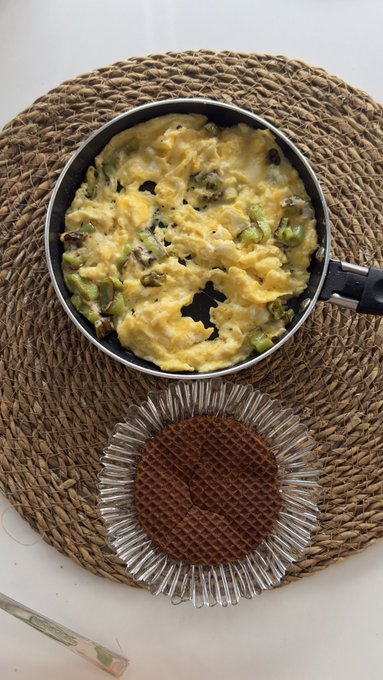 An overhead view of a pan containing scrambled eggs mixed with vegetables and mushrooms, placed next to a stroopwafel on a woven placemat.