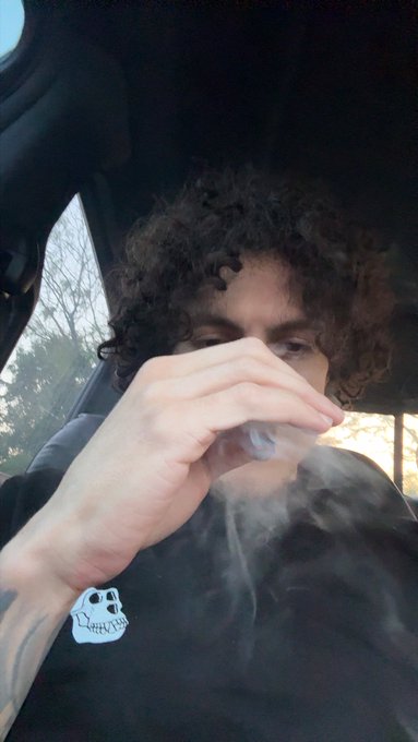 A person with curly hair sits in a car, holding a vape that produces a cloud of vapor, wearing a black t-shirt featuring a white skull graphic.