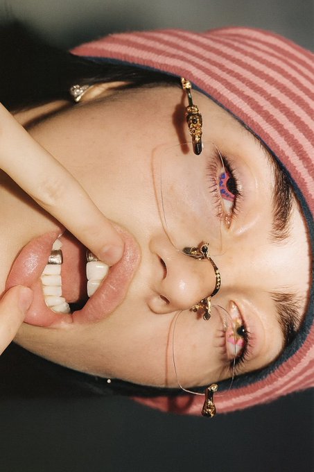 A close-up portrait shows a person wearing a striped pink bucket hat, gold facial jewelry, and dramatic eye makeup, with their fingers pulling their lip open to reveal teeth.
