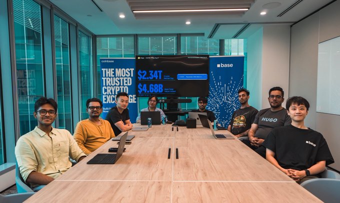 Team of seven developers seated around a conference table at Coinbase Base, with screens behind them displaying crypto exchange volume figures of $2.34T and $4.68B.