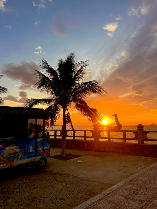 A vibrant sunset over a tropical beachfront with a palm tree silhouetted against orange and blue skies, featuring a colorful shaved ice cart labeled "JAMAICA SHAVED ICE" in the foreground. The serene seaside scene captures the peaceful, grateful vibe of the "Gm soapy fam • Enjoy every moment" morning greeting.