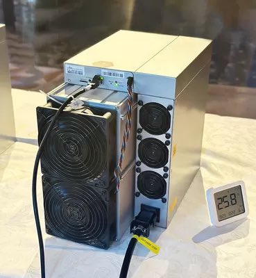 A cryptocurrency mining setup featuring stacked ASIC miners with cooling fans displayed on a table, alongside a digital thermometer reading 25.8.