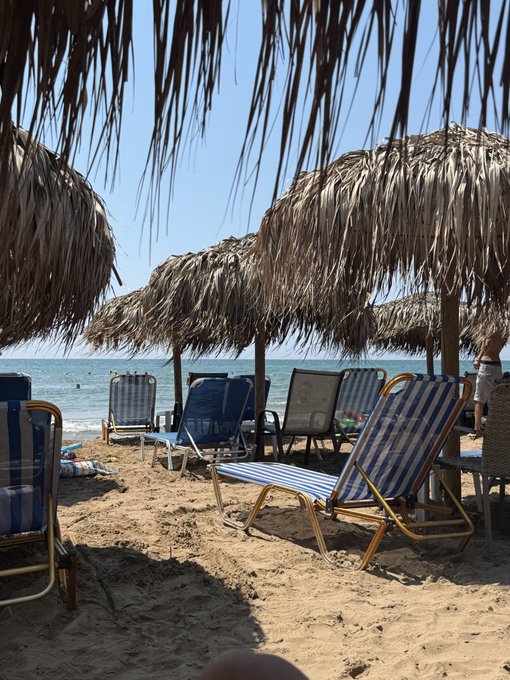 A sunny beach scene with rows of blue and white striped lounge chairs shaded by thatched straw umbrellas, with a clear blue ocean in the background.