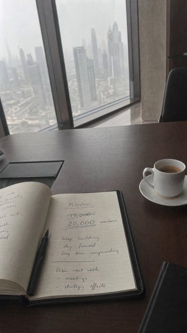 An open journal on a high-rise office table shows notes about 'Milestones' including '15,000 members' and '20,000 members', with a coffee cup and a city view in the background.