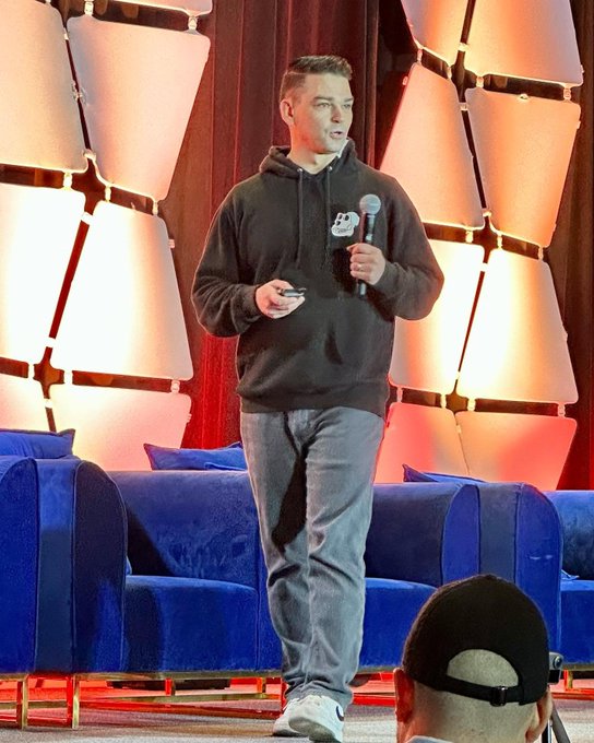 A young man in a black hoodie and gray jeans holds a microphone while speaking on a stage with blue couches and modern geometric wall panels behind him.