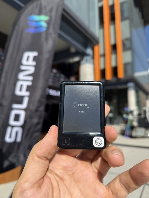 A hand holding a Ledger 'nibu' hardware wallet in front of a Solana banner at an outdoor event.