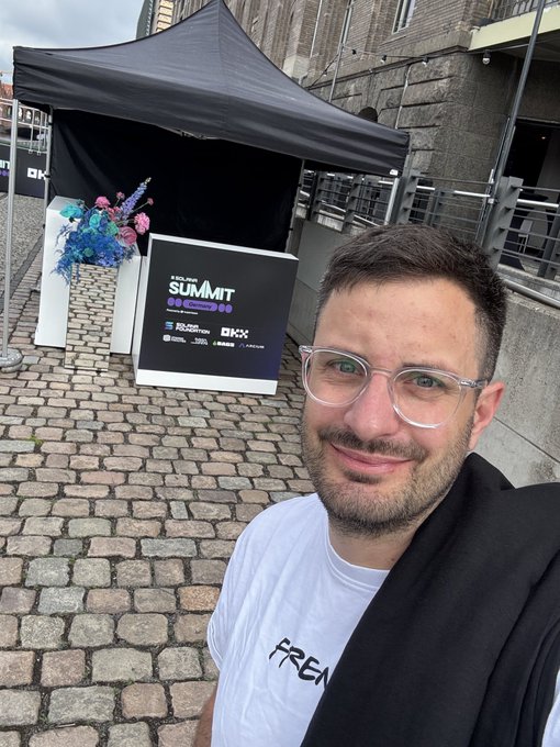 The image features a man wearing glasses standing outside of a tent or pavilion that has a black canopy. The man appears to be smiling, possibly posing for a photo. There are also some flowers visible near the tent, adding a touch of color to the scene. The text in the image reads "GM FRENS The day is finally here! The Solana Summit Germany kicks off in Berlin TODAY @FrensFactory is repping on the ground in Berlin to extract the best ecosystem alpha, network with builders, and bring the heat back to our holders Big things cooking today @SuperteamDE".