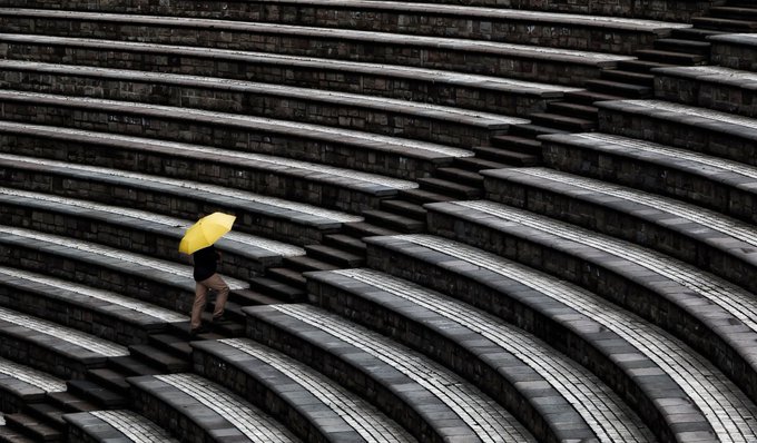 A photograph of a person holding a bright yellow umbrella walking up the gray stone steps of an outdoor amphitheater.
