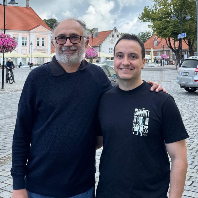 Two men posing for a photo in a European town square with cobblestone streets and historic buildings in the background.