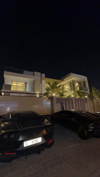 A sleek black sports car and a black sedan are parked in the driveway of a modern, illuminated luxury villa at night, with the sports car displaying a license plate reading "81282." The scene captures the glamorous nighttime vibe of the "new crib" referenced in the caption, with the lit-up architecture and high-end vehicles perfectly matching the boastful, celebratory tone of "Now we cook."