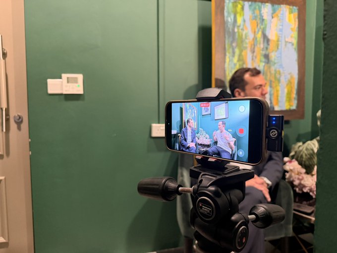 A smartphone mounted on a tripod is recording an interview or podcast between two men in a room with green walls and framed artwork, with the phone screen showing a recording interface.