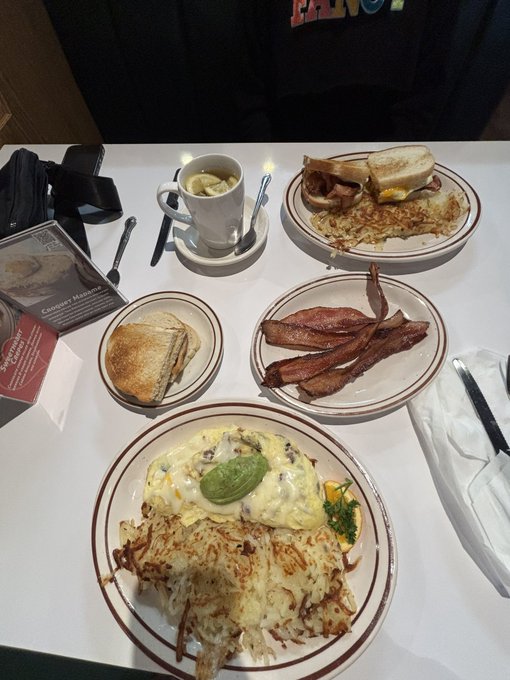 A diner-style meal on a table featuring plates of hash browns, bacon, eggs topped with avocado, a sandwich, bread, and a cup of coffee.