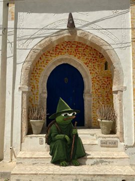 A Pepe the Frog figure dressed in a green wizard robe and pointed hat, sitting on steps in front of a doorway decorated with orange leaves, with a small sign reading 'SAX NOB'.