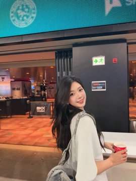 A young woman with long dark hair sits in a modern lobby or cafe area, turning to smile at the camera while holding a coffee cup.