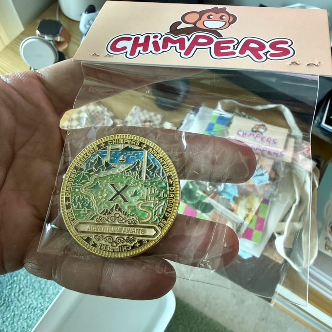 A hand holding a 'CHIMPERS' branded gold commemorative coin (reading 'ADVENTURE AWAITS' and 'CHIMPERS ADVENTURE') in its plastic packaging, with additional Chimpers merchandise visible in the background.