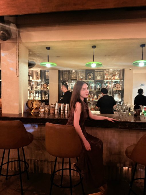 A young woman in an elegant burgundy dress poses gracefully at a sophisticated bar, illuminated by green pendant lights, with bartenders working behind the counter. The image captures the upscale nightlife atmosphere referenced in the post, reflecting the social event experience during her return visit to Bali.