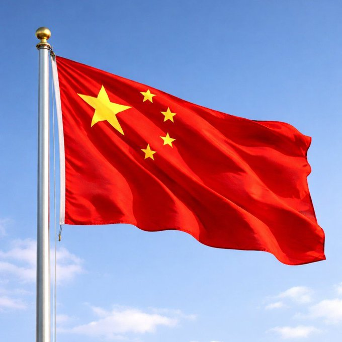 The image features a flag with red, yellow, and white colors flying high against a blue sky. The flag has three stars on its top half and four stars on its bottom half, representing the Chinese national flag. The flag is attached to a pole that stands tall in front of a backdrop of clouds.