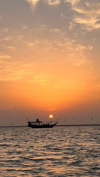 A silhouetted boat carrying several people floats on water against a vibrant orange sunset sky dotted with flying birds.
