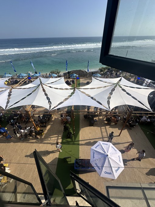 An elevated view of an outdoor beachside venue featuring white tent canopies, tables, and a large white 'Dash' umbrella on artificial turf overlooking the ocean.