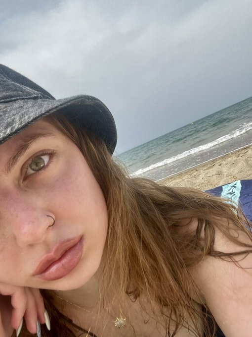 A close-up selfie of a young woman with long hair, a nose ring, and a black cap at the beach, with ocean waves and sand visible behind her.