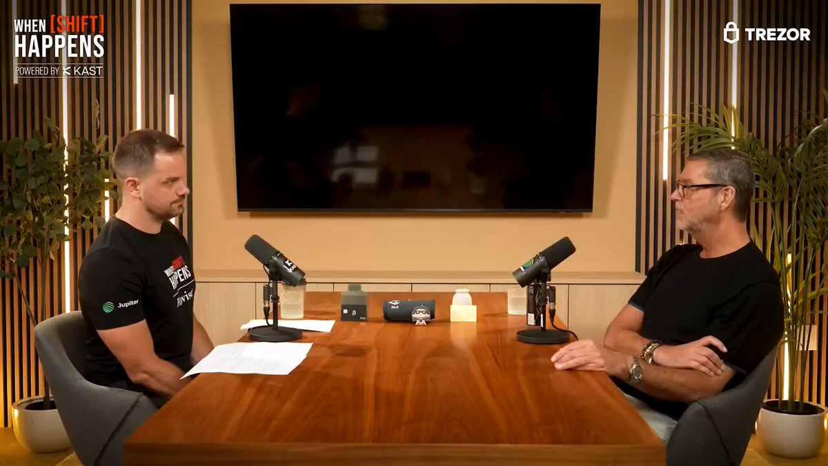 The image shows two men sitting at a table having a conversation. They are wearing black shirts and appear to be engaged in a discussion or interview, possibly related to cryptocurrency. The background features a television screen displaying an advertisement for Bitcoin Cash, which is likely related to their conversation.