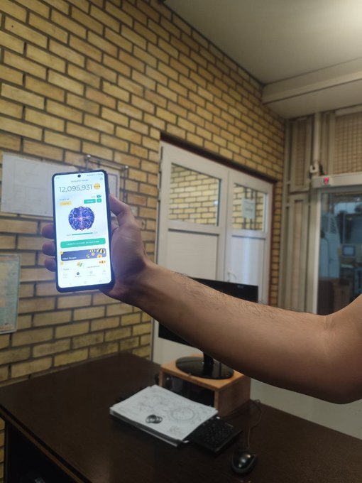 A person's hand holding up a smartphone displaying an app with the number '12,095,931' on the screen, in what appears to be an office or workshop setting.