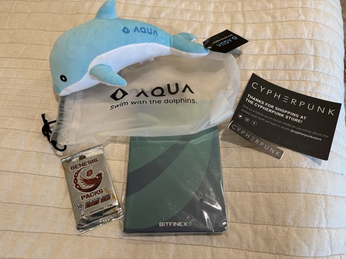 A haul from Cypherpunk Store includes a blue "Aqua - Swim with the dolphins" plush dolphin, a Bitfinex-branded notebook in plastic wrap, a "Genesis Packs" pack of 10 historical crypto events cards, and a Cypherpunk-branded card thanking the customer for shopping—aligning with the post celebrating Bitcoin-business purchases and the dolphin's Bitcoin association.