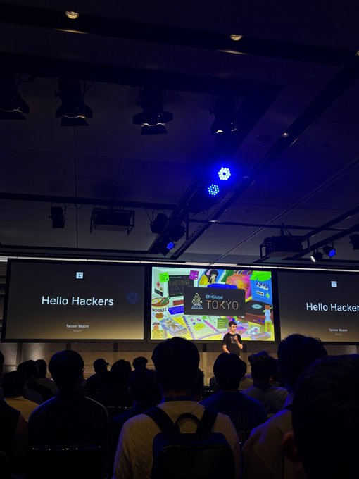 The image shows a presentation at ETHGlobal Tokyo, where Tanner Moore stands on stage in front of screens displaying "Hello Hackers" and the event logo, addressing an audience of attendees seated in a dimly lit conference hall.