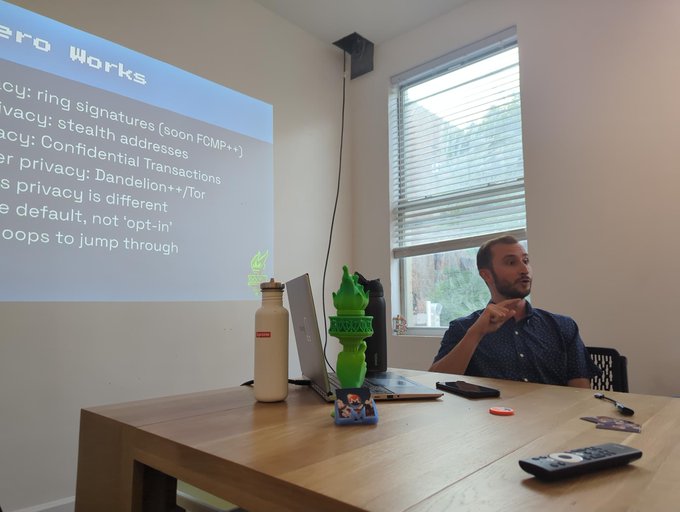 A man from Cake Wallet sits at a table presenting a slide titled "How Monero Works," which lists features like ring signatures (soon FCMP++), stealth addresses, Confidential Transactions, and Dandelion++/Tor, during the first Monero Meetup in NYC.