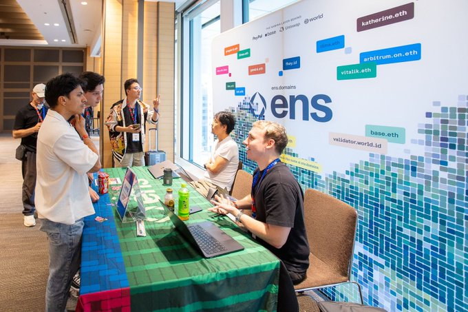 A conference or networking event where attendees gather at an 'ens' (Ethereum Name Service) booth, featuring branding for various .eth domains like katherine.eth, arbitrum.on.eth, and vitalik.eth.