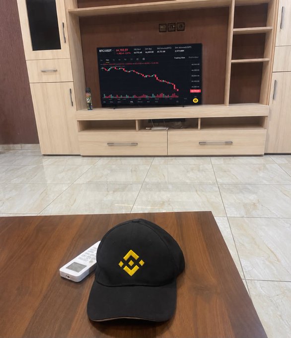 A black cap featuring the yellow Binance (BNB) logo rests on a wooden table next to a TV displaying a BTC/USDT trading chart with red price movements.
