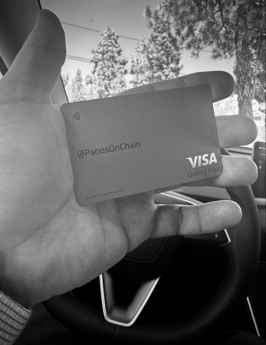 A hand holds a Visa Debit card printed with '@PanosOnChain' inside a car, shown in black and white.