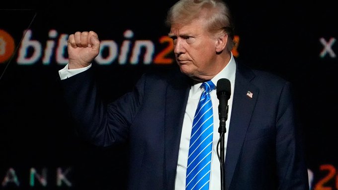 Donald Trump raises his fist while speaking at a podium at the Bitcoin 2024 conference, with the event's logo visible behind him. The image relates to the post's discussion of Trump's financial interests in cryptocurrency, as he has positioned himself as a pro-crypto candidate while Democrats push for conflict-of-interest provisions in the CLARITY Act to address his crypto holdings.