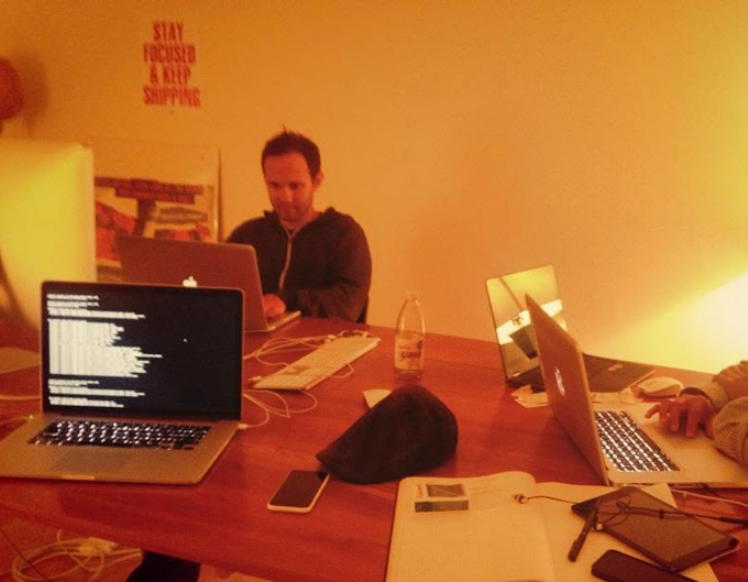 Several people work on laptops at a wooden table in a warmly lit room, with a poster on the wall reading 'STAY FOCUSED & KEEP SHIPPING'.