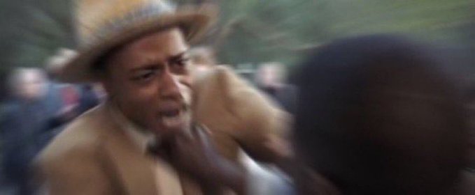 A low-resolution action shot showing a man wearing a straw hat and tan jacket appearing to shout or yell intensely during what looks like a chaotic crowd scene.