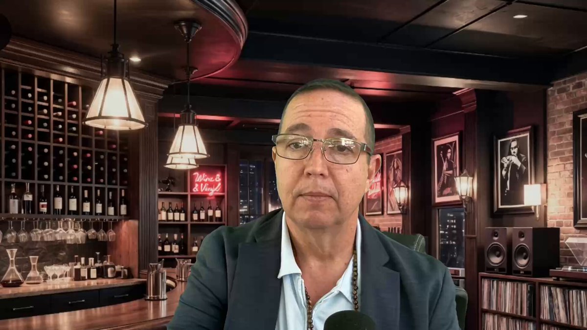 The image features a man sitting at a bar, wearing glasses and a suit jacket. The background of the scene includes wine bottles displayed behind him, suggesting that he might be in a winery or a bar with a wine selection. There are also several books visible on shelves nearby, indicating an intellectual atmosphere.