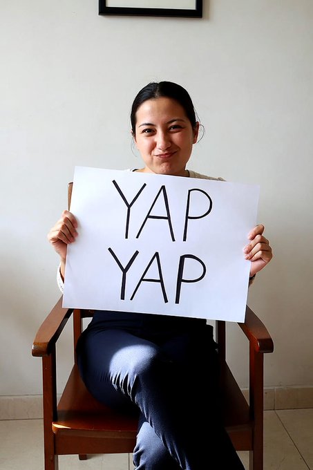 A woman sits in a wooden chair smiling while holding up a sign that reads 'YAP YAP' in bold black letters.