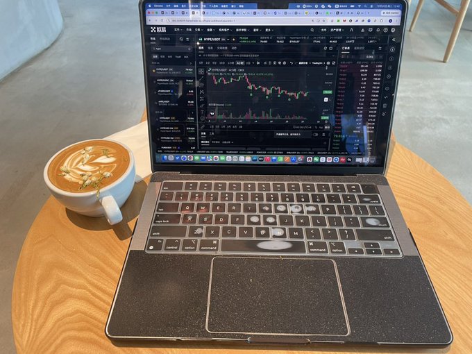 A laptop sitting on a wooden table displays cryptocurrency charts on the OKX exchange interface, accompanied by a cup of latte art coffee.