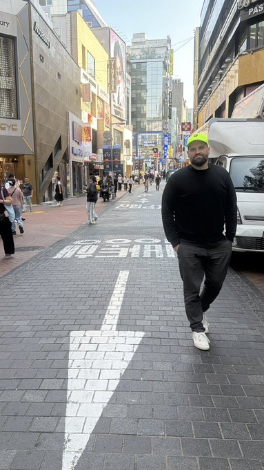 A bearded man in a black sweater, gray pants, and a neon yellow cap stands on a busy city street, possibly in Tokyo given the Japanese signage in the background.