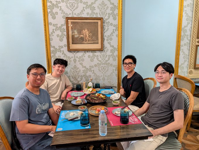 Four young men sitting around a dining table with plates of food and water bottles, in a room with patterned wallpaper and a framed picture.